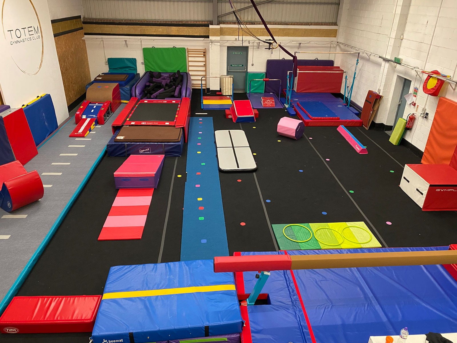 A vibrant photograph of children joyfully participating in a gymnastics class at Totem Gymnastics, showcasing the equipment like bars, vault, foam pit, beam and floor. diverse group of kids engaged in various activities. Features the gym's colourful and safe environment.
